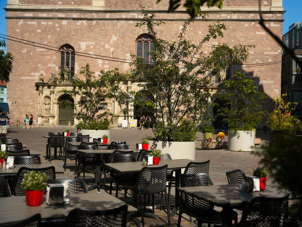 pilsētas ainava ar vēsturiskas ēkas fasādi, kur redzami arku logi un koki priekšplānā